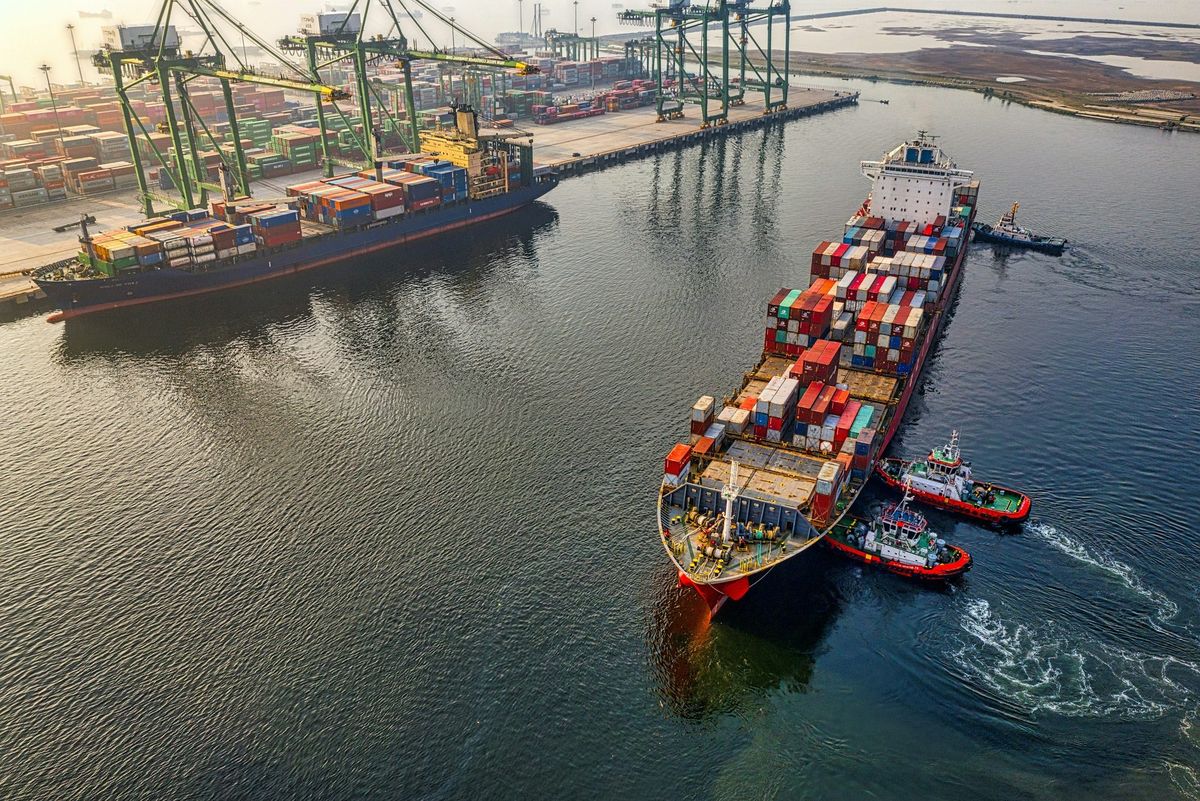 Containerschiff mit Schleppern neben einem Containerterminal mit Containerbrücken