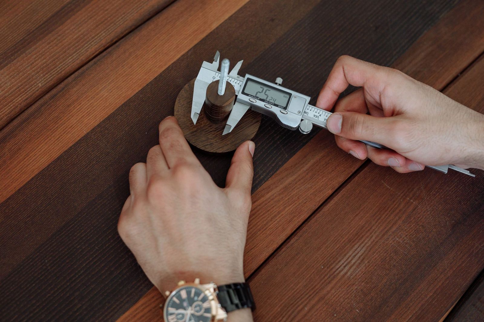 Hands measuring a turned wooden part with a digital caliper on a wooden table