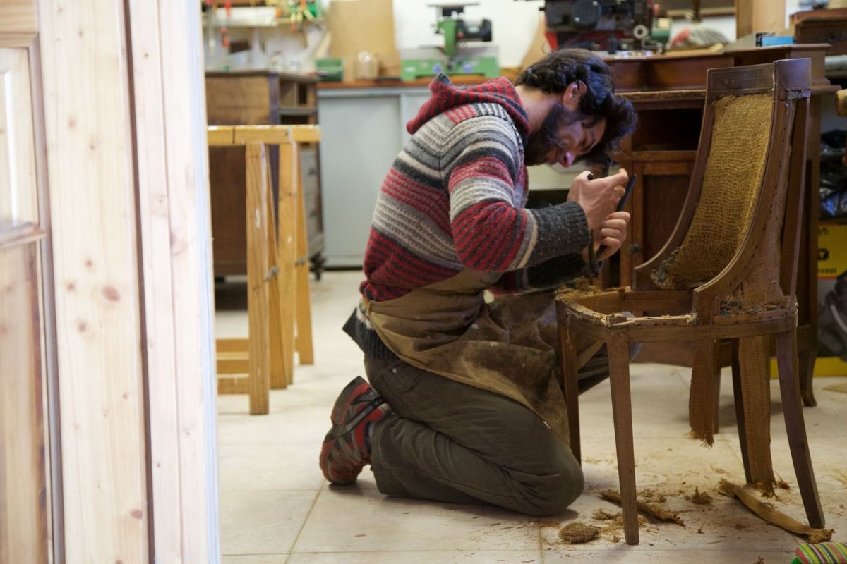 Meșter îngenuncheat într-un atelier, care îndepărtează tapițeria veche de pe scheletul unui scaun din lemn