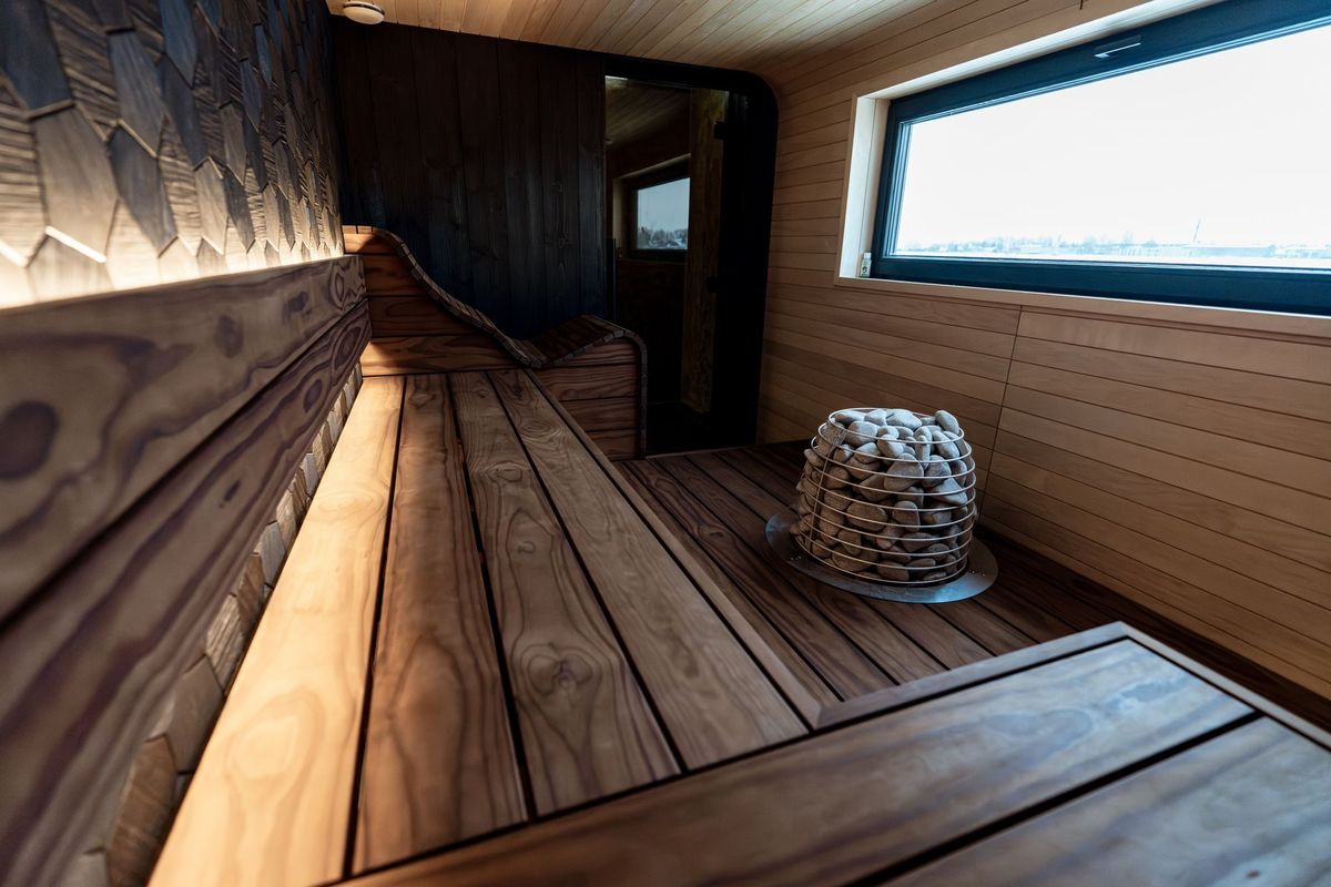 Sauna with dark-brown wooden benches and floorboards, a stone heater and a long window