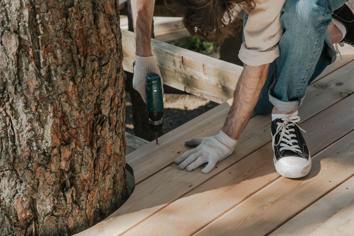 Gloved hands screwing down new deck boards with a cordless drill next to a tree trunk