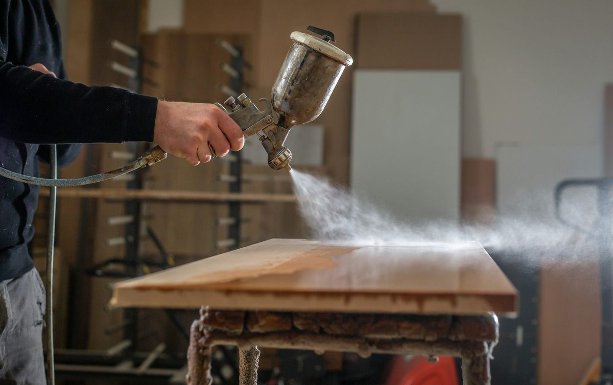 Werker spritzt in der Werkstatt mit einer Lackierpistole Lack auf eine Massivholz-Tischplatte
